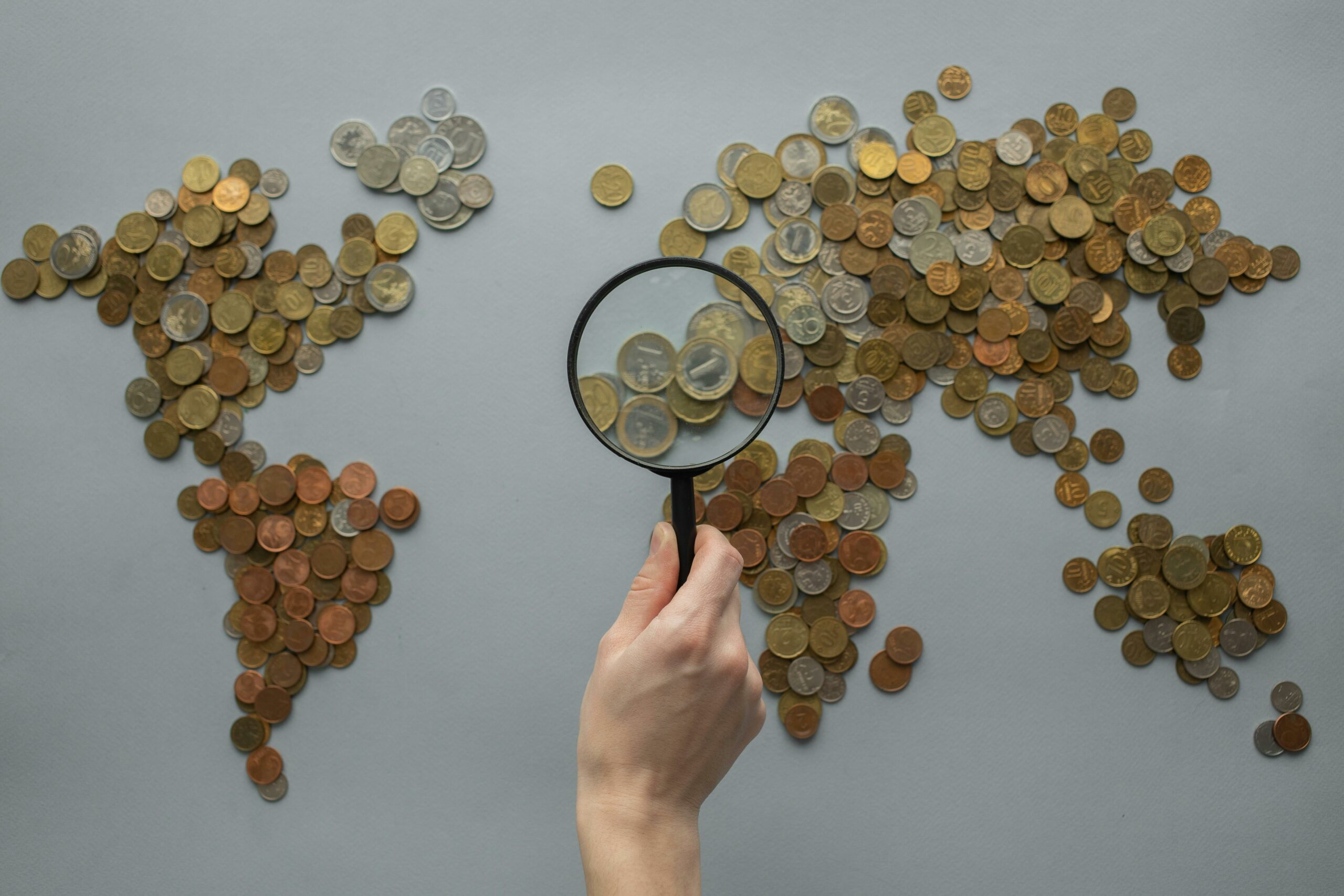 COINS LAID OUT IN SHAPE ON WORLD MAP, PERSON HOLDING MANGIFYING GLASS